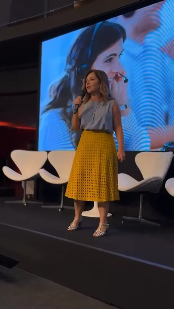 Claudia Fernandes de corpo inteiro num palco grande, microfone na mão, imagem dela no telão ao fundo